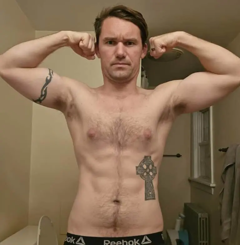A man stands in a bathroom flexing his biceps, tattoos visible on his left arm and lower abdomen, showcasing the results of dedicated training with guidance from an online fitness coach.